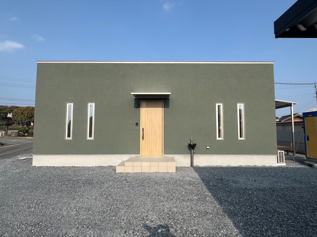 Front exterior of a minimalist gray building with a wooden door and four tall narrow windows, set on gravel under a clear blue sky.
