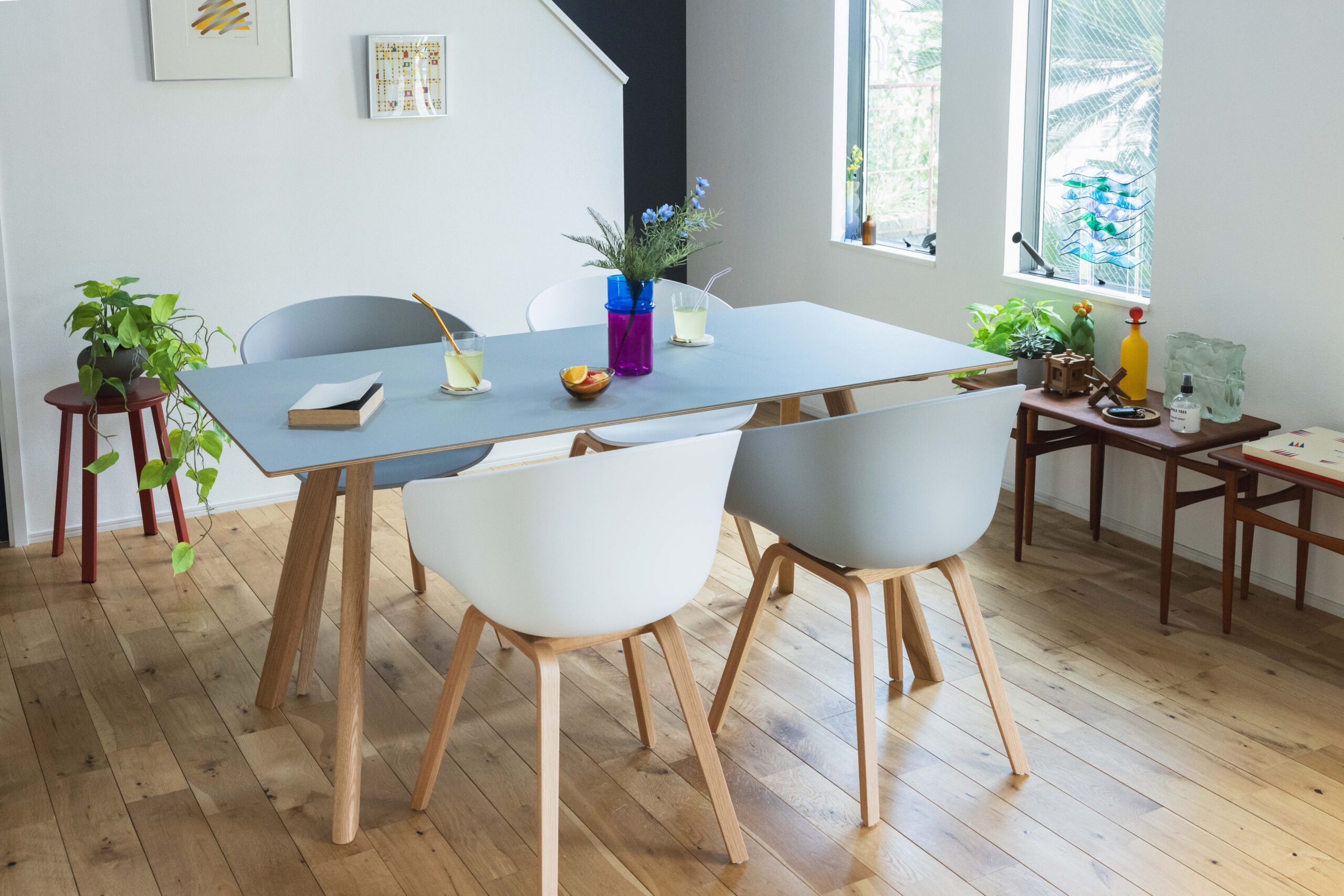 Bright dining area with a pale blue table and white chairs, wooden legs, and houseplants by a sunlit windowed wall.