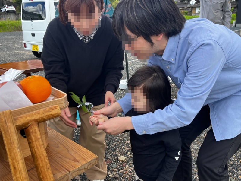 Two adults help a child hold a small leafy branch outdoors, with an orange resting on a wooden stand nearby.