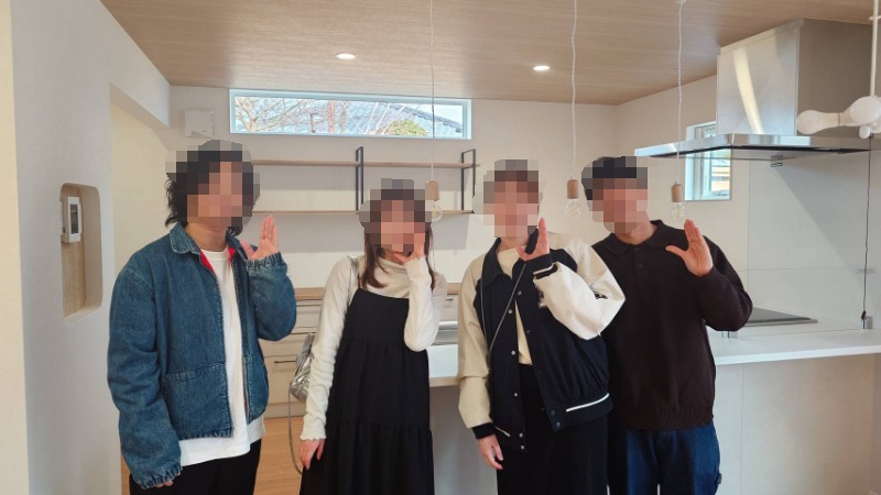 Four friends posing in a bright, modern kitchen, making peace signs at the camera (faces blurred). Those on the left wear a denim jacket and dress; others wear casual outfits.