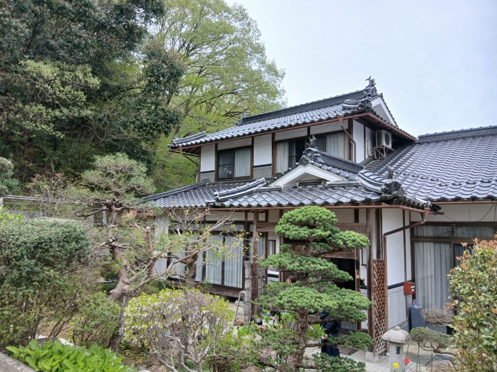 Traditional tiled-roof house with wooden lattice, surrounded by a lush garden and trimmed bonsai trees by a sunny yard