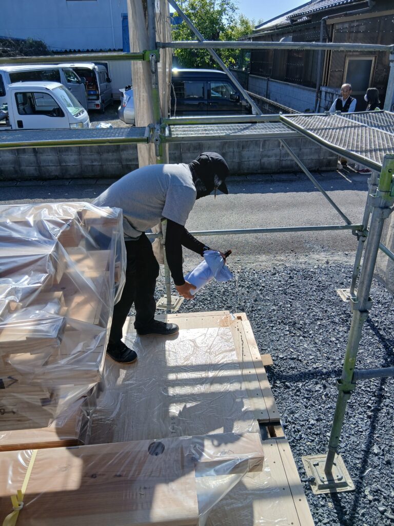 Construction worker in a gray shirt bends over to inspect a plastic‑wrapped pallet of lumber on a scaffolded work area outside.