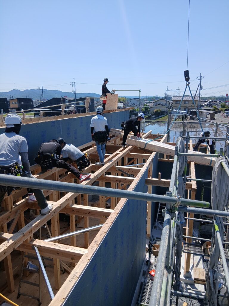 Construction workers assemble wooden framing on a building's second floor, wearing hard hats and safety gear.