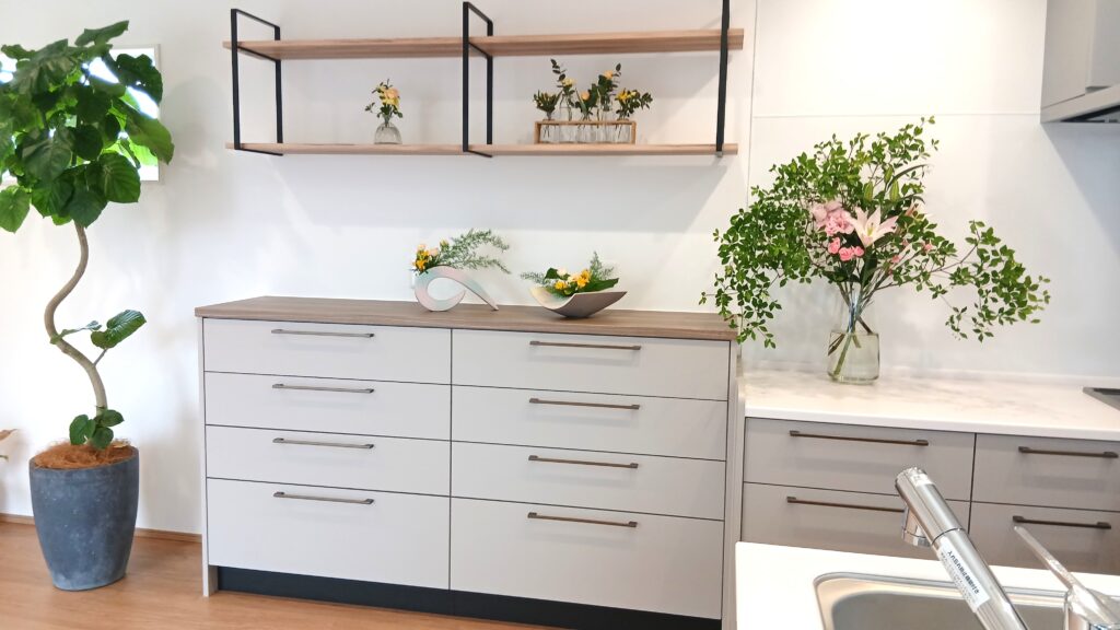 Modern kitchen scene with light gray drawers, wooden countertops, and open wood-and-metal shelves, accented by potted plants and vases with flowers.