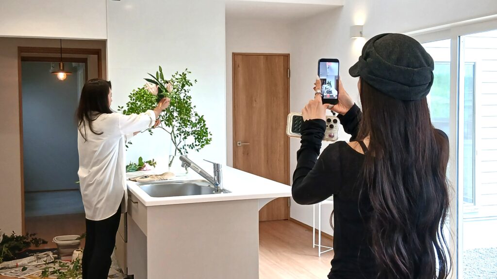 Two women in a bright kitchen: one arranging a flower bouquet at a counter while the other shoots a photo with a smartphone.