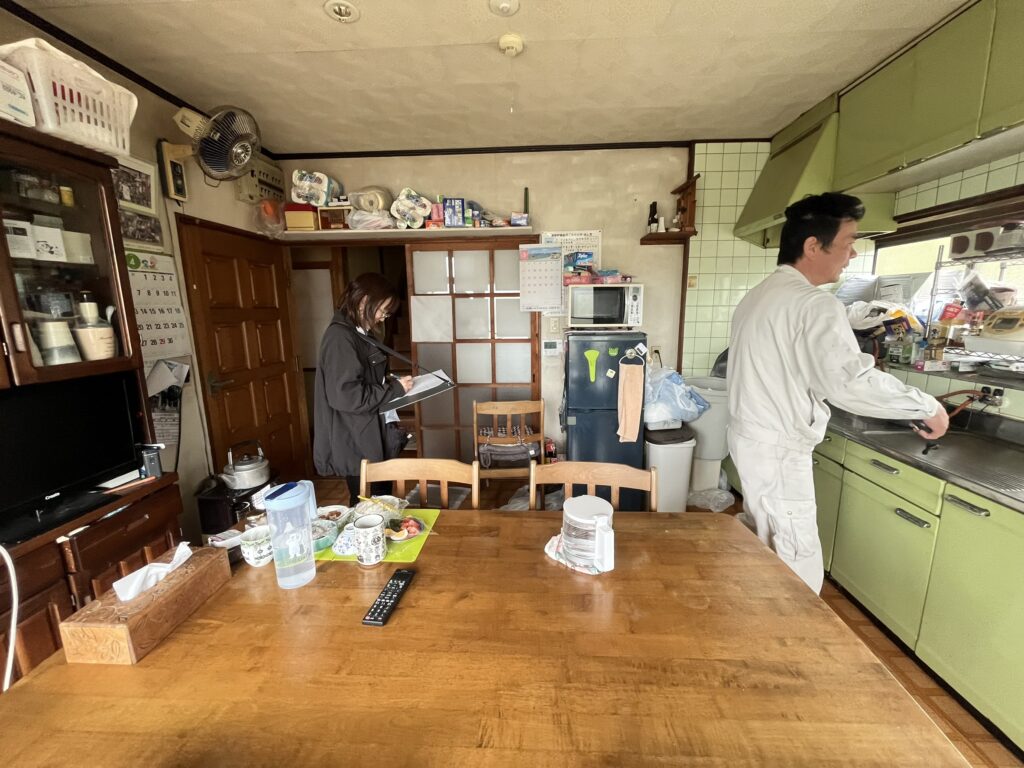 Kitchen/dining area with a long wooden table; a woman takes notes in the background while a man in a white uniform works at a counter to the right; cluttered shelves, cups, and a water bottle on the table.