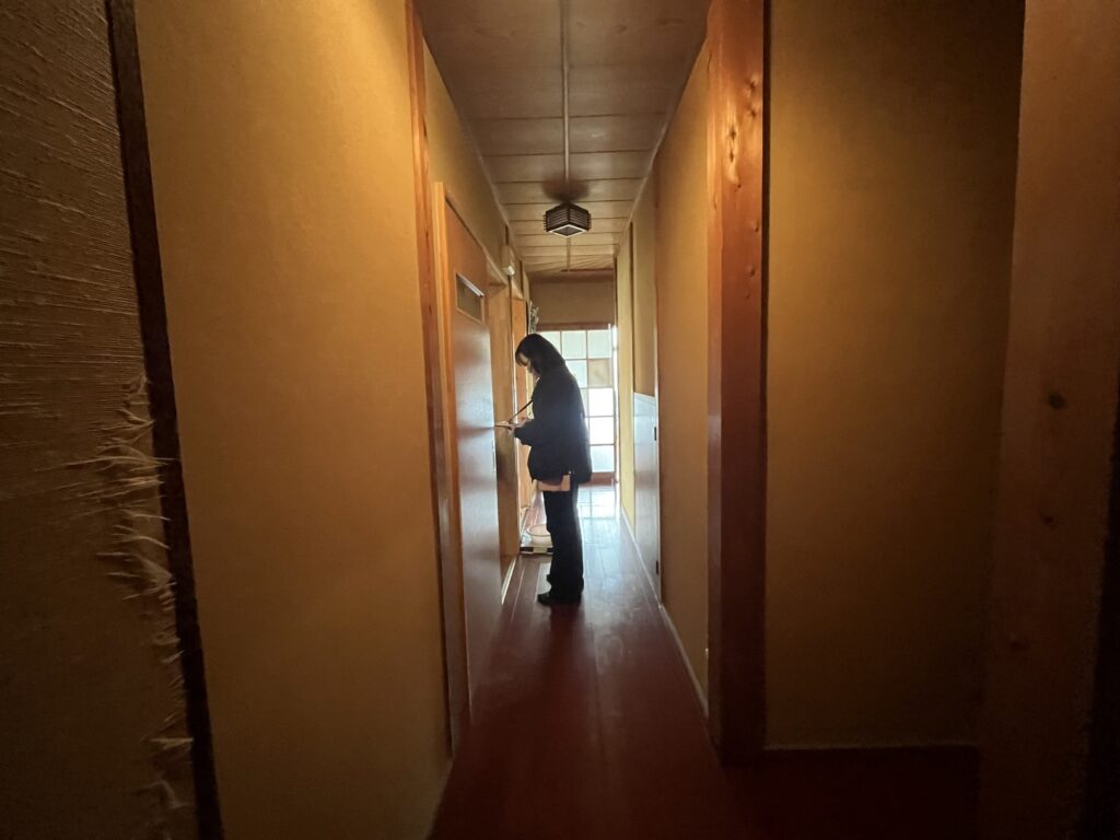 Person standing in a dim, narrow wooden hallway, looking down at a device in their hands near a window at the far end.