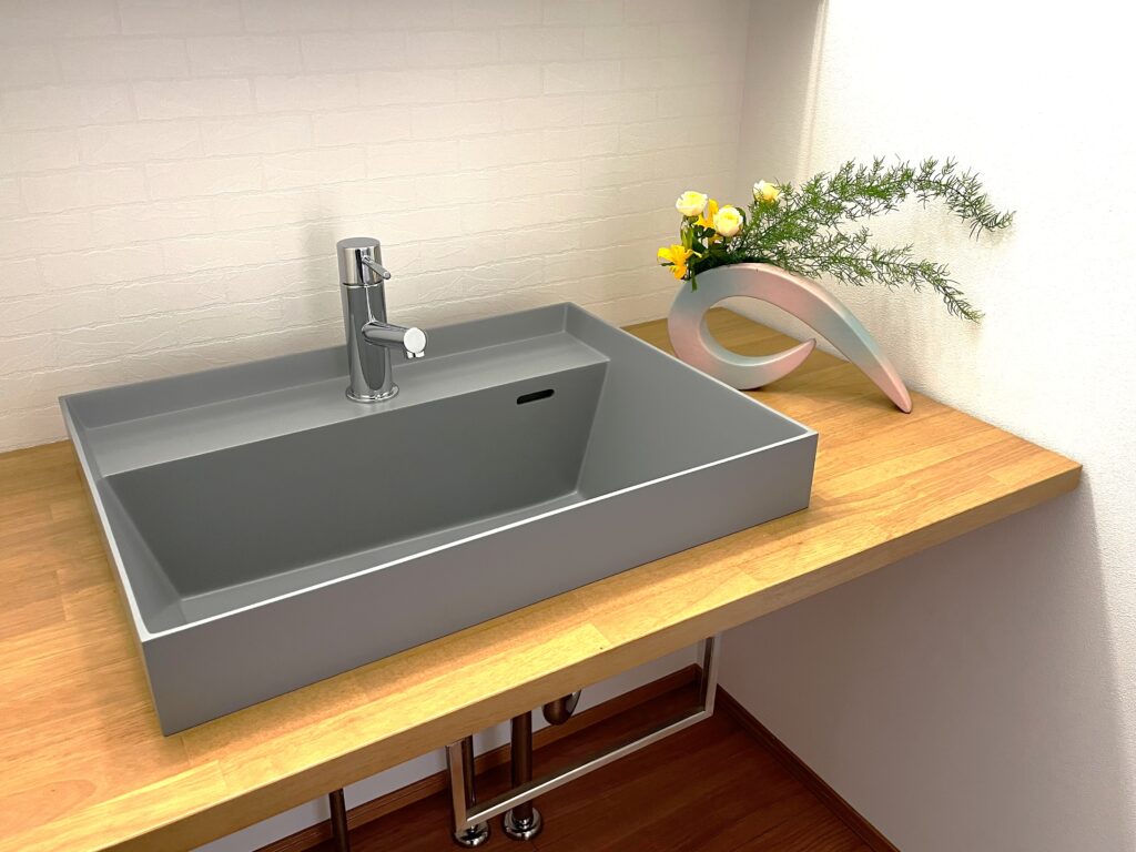 Modern gray rectangular bathroom sink with chrome faucet on a wooden counter, next to a curved decorative vase with yellow and white flowers.