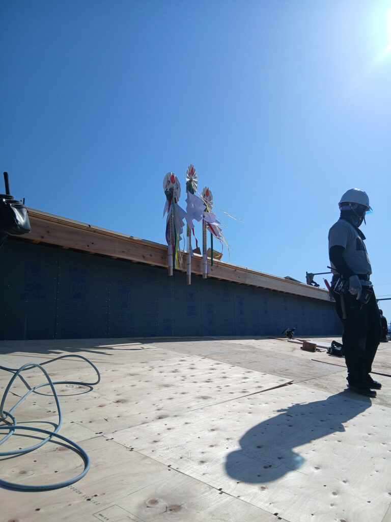 Construction worker in a hard hat standing on a roof edge with colorful ribbon windsocks, under a clear blue sky near solar glare from the sun.