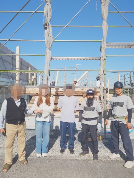 Six people standing in front of a building under construction with scaffolding, wearing helmets and safety gear, posing for a photo.