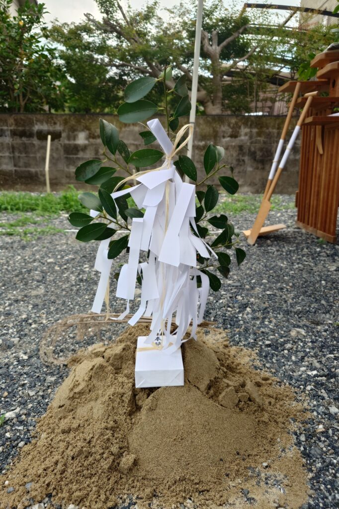 Small leafy branch tied with white ritual paper strips (shide) on a sandy mound, in a gravel yard setting, likely a Shinto shrine tree ceremony