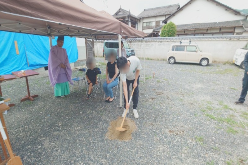 Ground-breaking ceremony under a canopy; a priest in purple robes watches as a person shovels sand while others sit nearby.