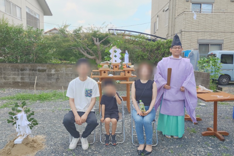 Family of four seated for a Shinto ceremony outdoors, with a priest in purple robes beside a ceremonial altar in the background.